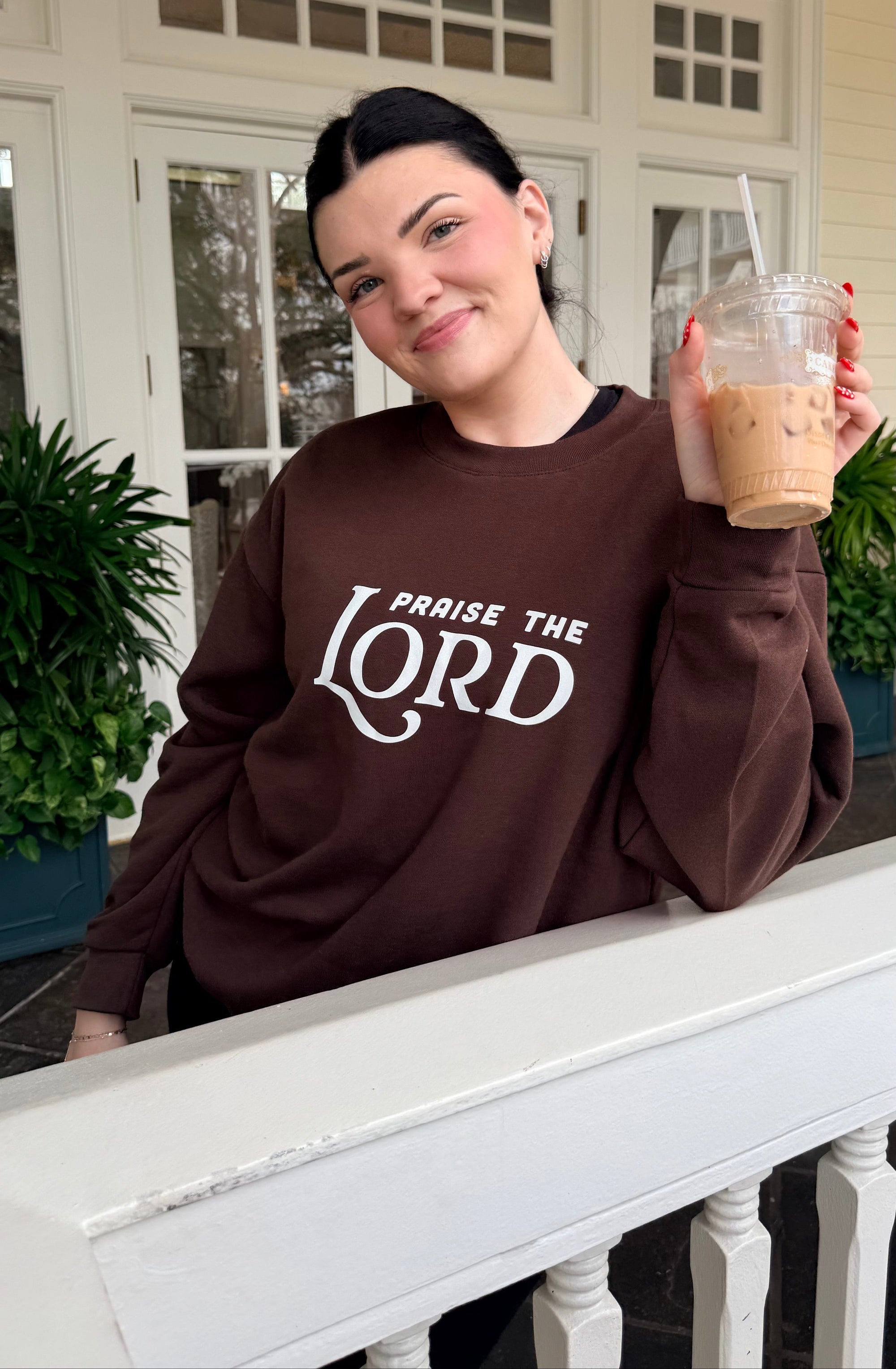 Person wearing a brown sweatshirt with 'Praise the Lord' text, holding a drink on a porch.