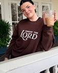 Person wearing a brown sweatshirt with 'Praise the Lord' text, holding a drink on a porch.