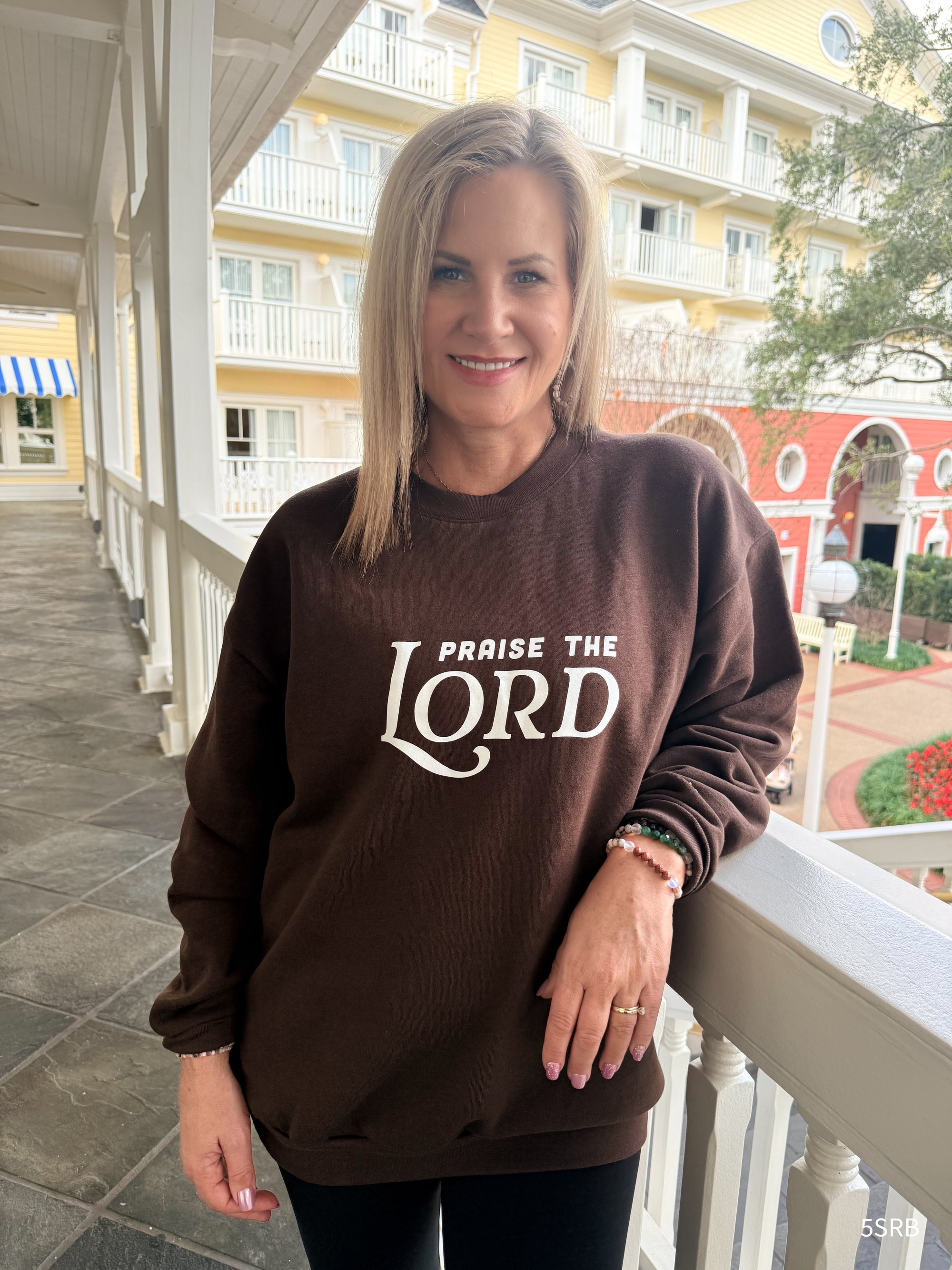 Woman wearing a brown sweatshirt with 'PRAISE THE LORD' text on a porch.