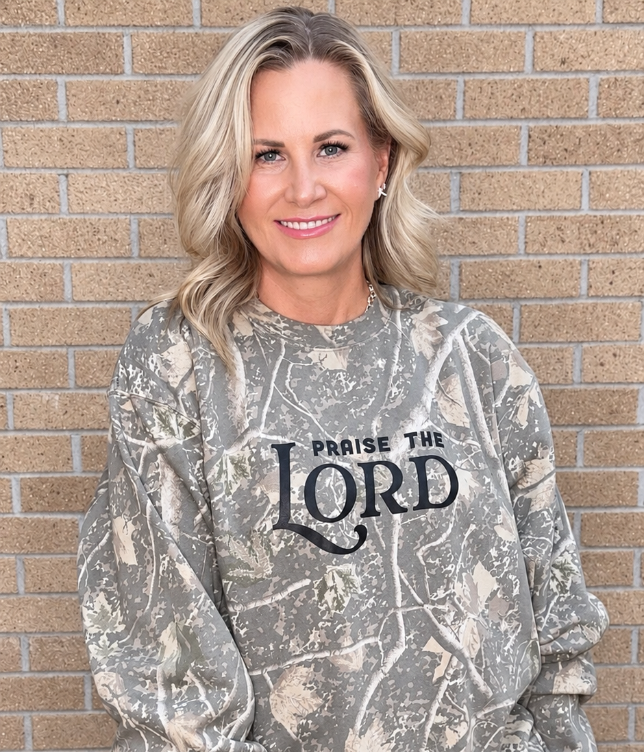 A woman wearing a long-sleeve crewneck sweatshirt with a brown and beige camouflage pattern and 'PRAISE THE LORD' printed in black across the chest, paired with light blue jeans, standing against a tan brick wall.