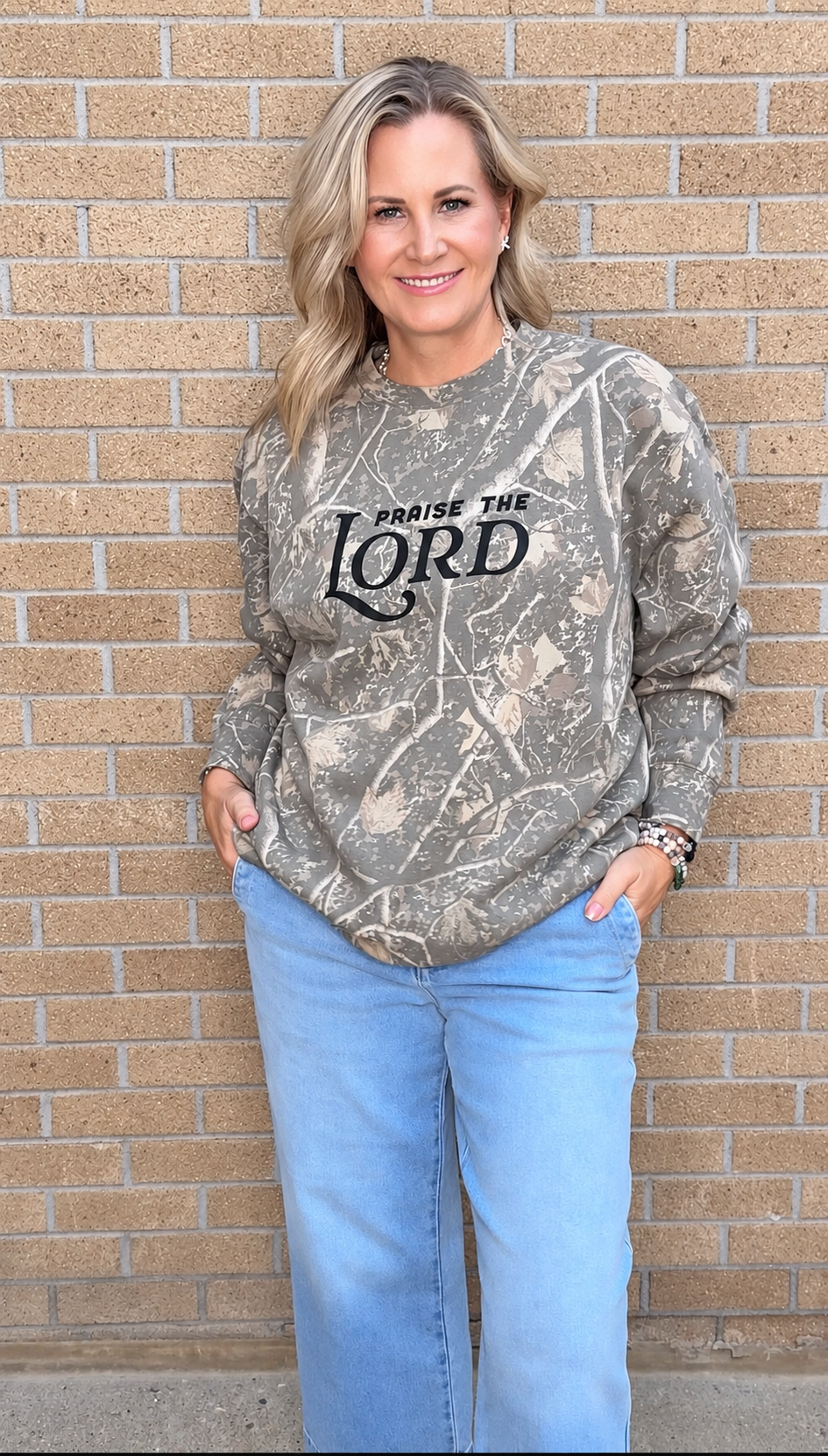 A woman wearing a long-sleeve camouflage sweatshirt with 'PRAISE THE LORD' printed in black across the chest, paired with light blue jeans, standing against a tan brick wall.