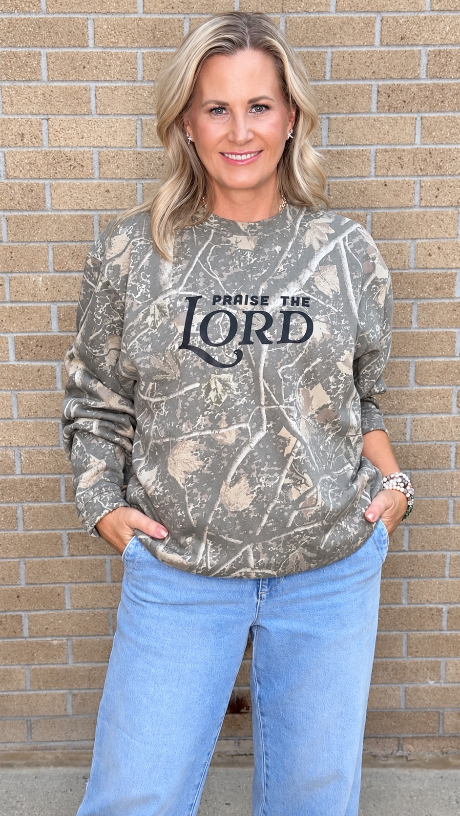 A woman wearing a camouflage-patterned sweatshirt with 'PRAISE THE LORD' printed in black across the chest, paired with light blue jeans, standing against a tan brick wall.