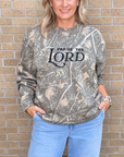 A woman wearing a camouflage-patterned sweatshirt with 'PRAISE THE LORD' printed in black across the chest, paired with light blue jeans, standing against a tan brick wall.