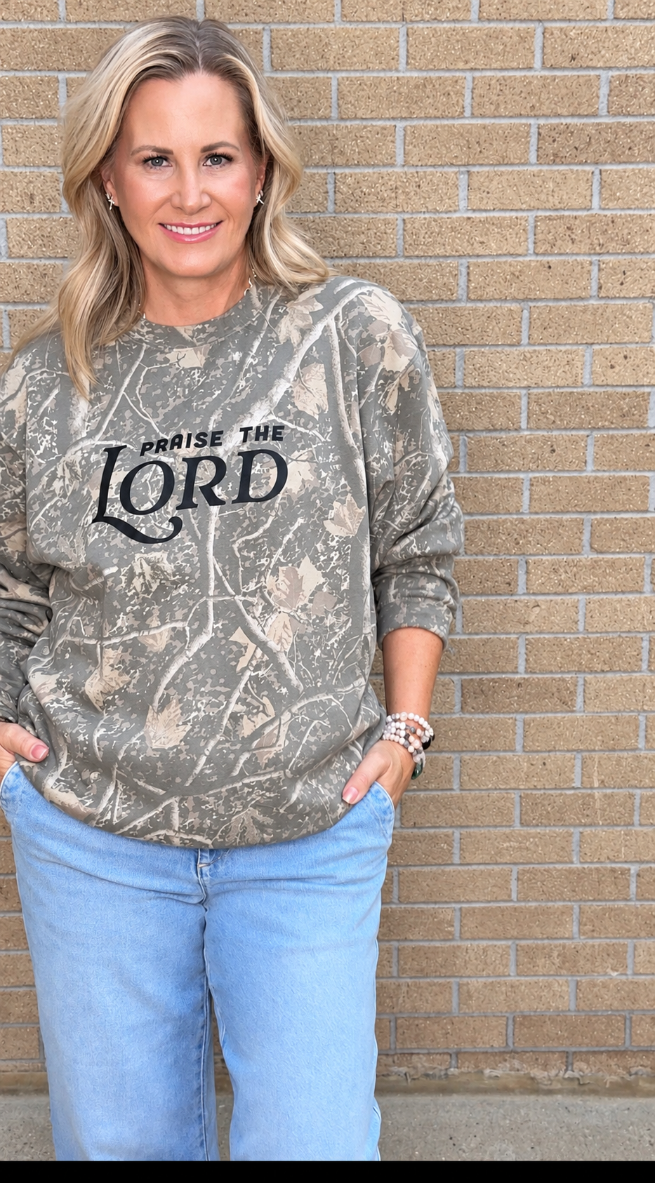 A woman wearing a camouflage crewneck sweatshirt with 'PRaise THE LORD' printed in black text, paired with light blue jeans, standing against a tan brick wall.