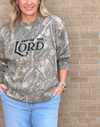 A woman wearing a camouflage crewneck sweatshirt with 'PRaise THE LORD' printed in black text, paired with light blue jeans, standing against a tan brick wall.