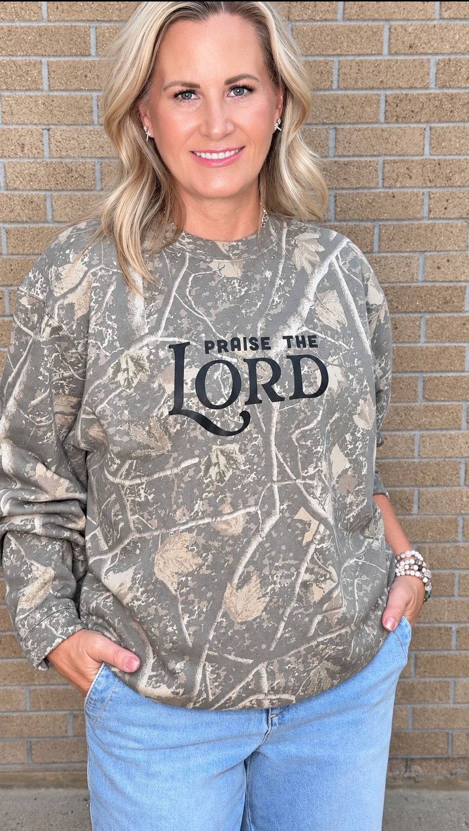 A woman wearing a camouflage-patterned long-sleeve sweatshirt with 'PRaise THE LORD' printed in black text on the front, paired with light blue jeans, standing against a tan brick wall.