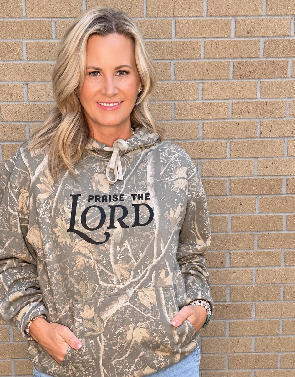 A woman wearing a camouflage hoodie with 'PRAISE THE LORD' printed on the front, paired with light blue jeans, standing against a tan brick wall.