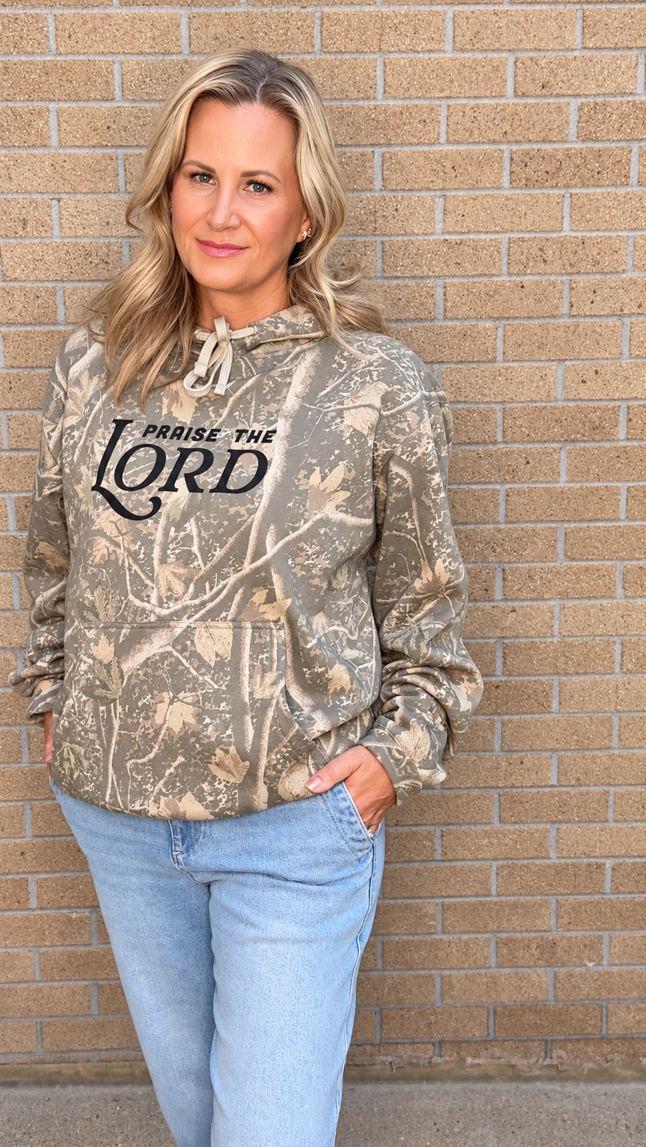 A woman wearing a camouflage hoodie with 'PRaise THE LORD' printed in black across the chest, paired with light blue jeans, standing against a tan brick wall.