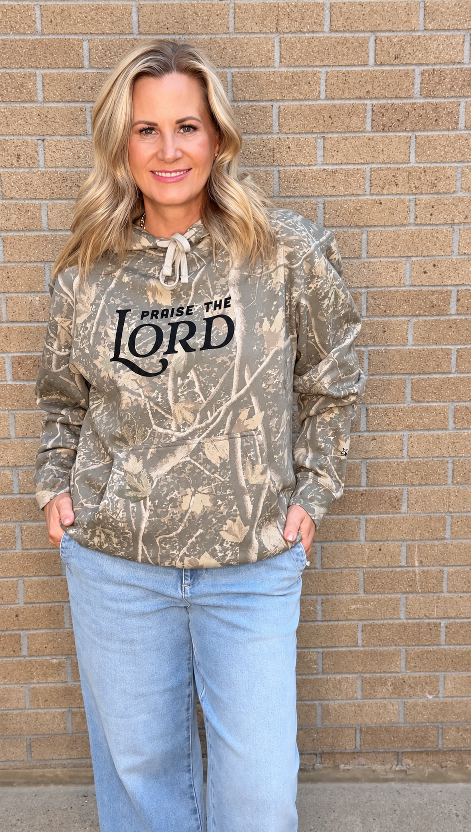 A woman wearing a camouflage hoodie with 'PRaise THE LORD' printed in black across the chest, paired with light blue jeans, standing against a tan brick wall.