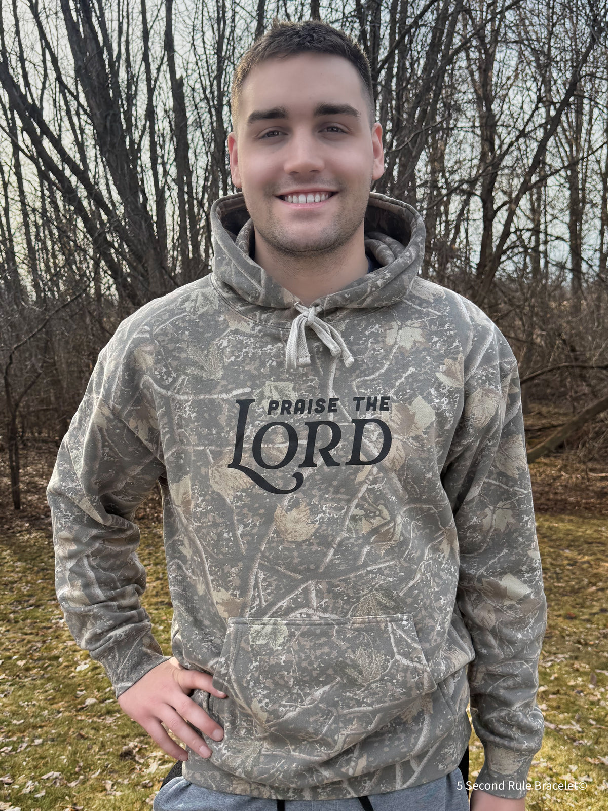 Person wearing a camouflage hoodie with 'Praise the Lord' text outdoors.