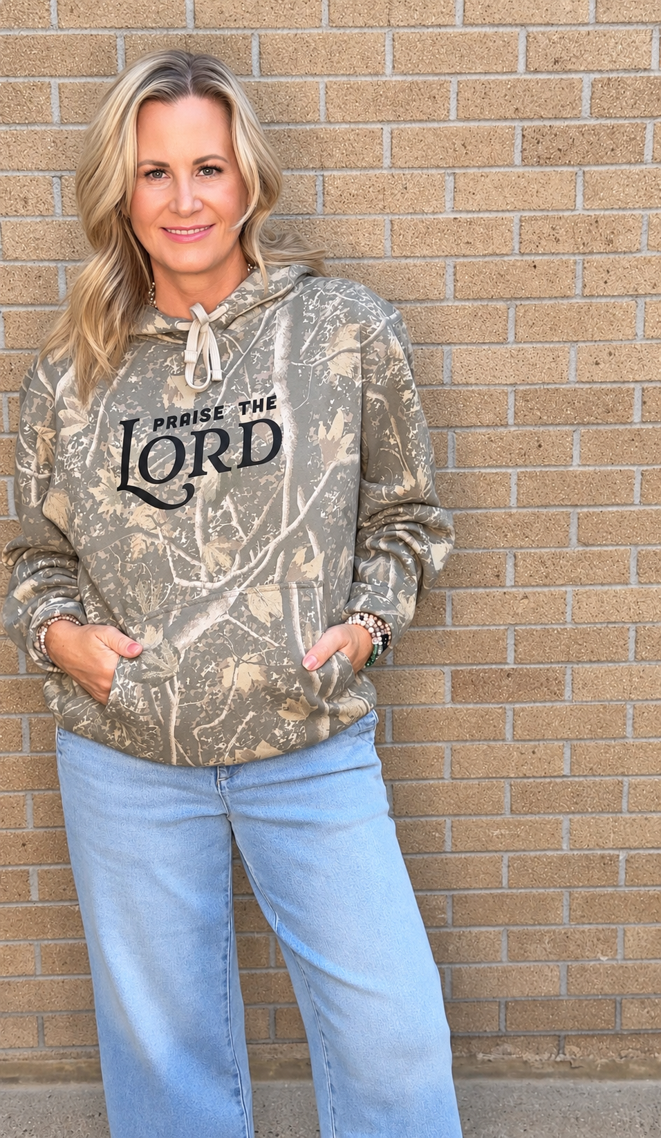 A woman wearing a camouflage-patterned hoodie with 'PRaise THE LORD' printed on the front, paired with light blue jeans, standing against a tan brick wall.