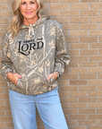 A woman wearing a camouflage-patterned hoodie with 'PRaise THE LORD' printed on the front, paired with light blue jeans, standing against a tan brick wall.