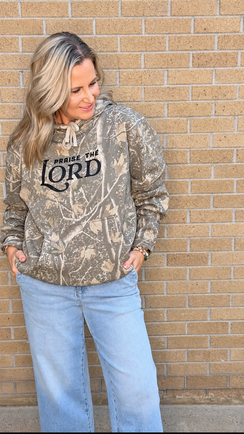 A woman wearing a camouflage hoodie with 'PRaise THE LORD' printed in black on the chest, paired with light blue jeans, standing against a tan brick wall.