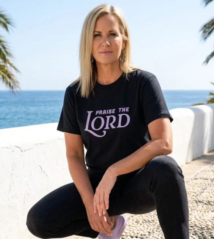 Woman wearing a black t-shirt with 'Praise the Lord' text, sitting by a coastal area with palm trees.