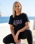 Woman wearing a black t-shirt with 'Praise the Lord' text, sitting by a coastal area with palm trees.