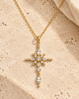 A gold cross pendant necklace adorned with clear gemstones, displayed on a textured beige stone surface.