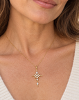 A woman wearing a delicate gold cross pendant necklace with small clear stones and a central pearl-like drop, styled with a white knit V-neck top.