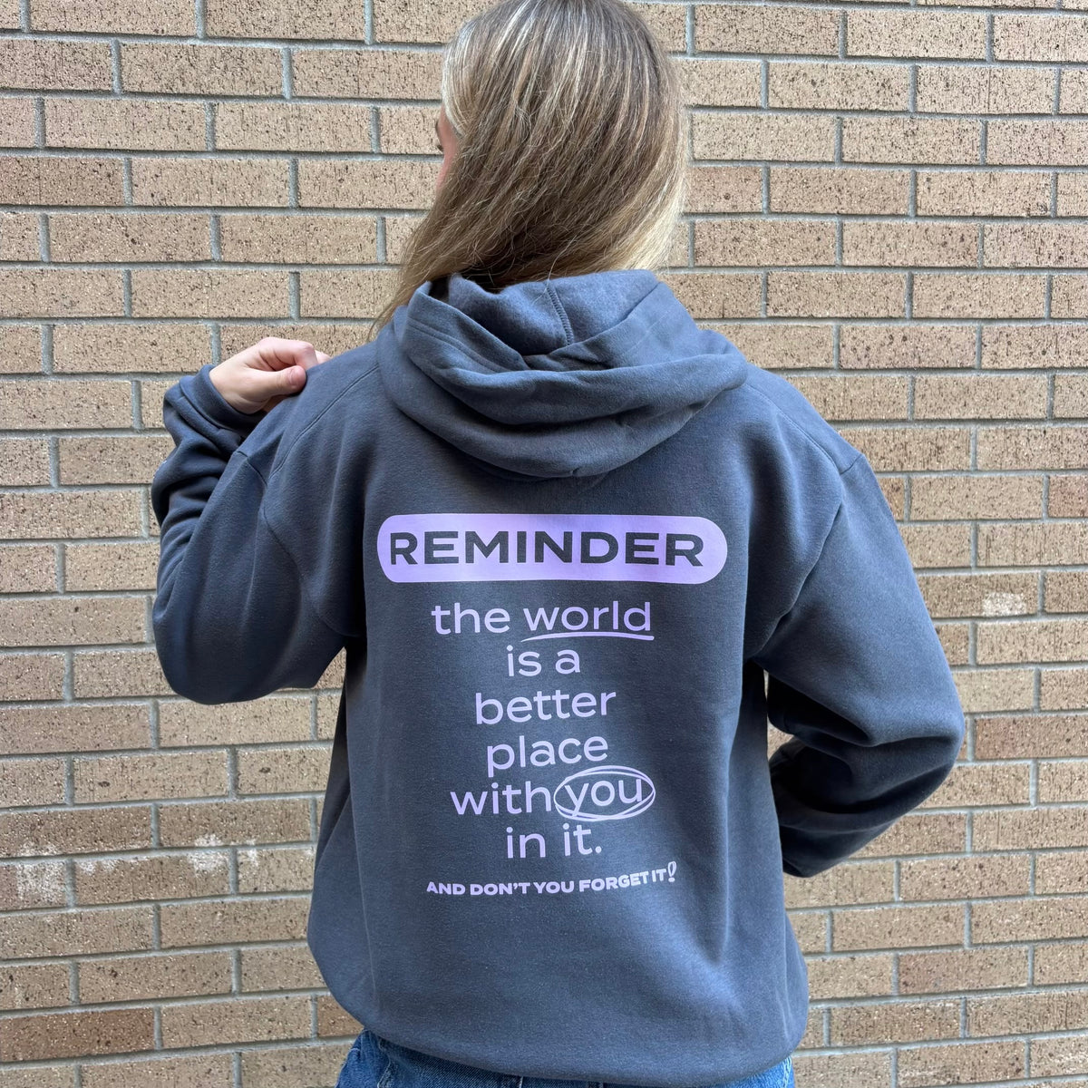 A woman viewed from behind wearing a slatedark gray hoodie with a motivational quote printed on the back, standing against a brick wall.