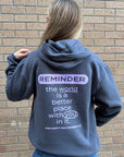 A woman viewed from behind wearing a slatedark gray hoodie with a motivational quote printed on the back, standing against a brick wall.
