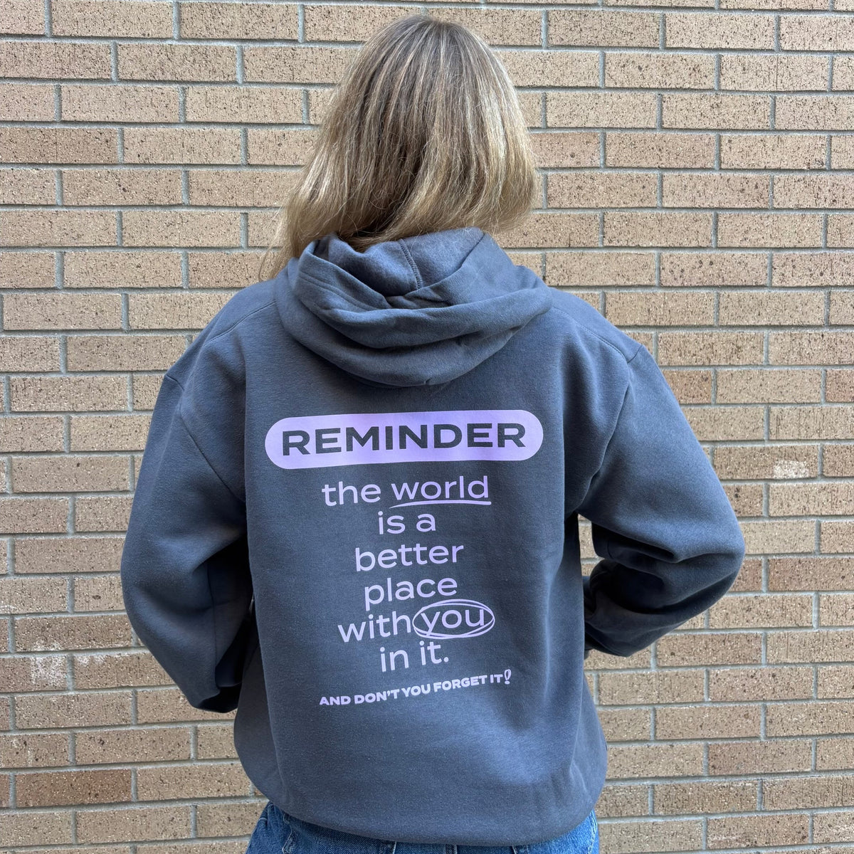 A person viewed from behind wearing a slate blue hoodie with a purple and white graphic text on the back, standing against a tan brick wall.