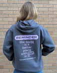A person viewed from behind wearing a slate blue hoodie with a purple and white graphic text on the back, standing against a tan brick wall.