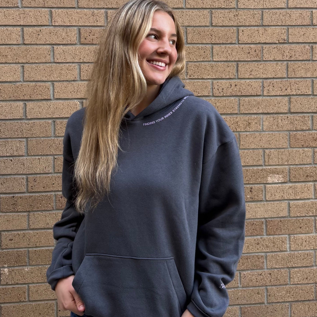 A woman with long blonde hair smiling and looking to the side, wearing a dark charcoal gray oversized hoodie and blue jeans, standing in front of a brown brick wall.