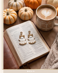 A pair of white ghost-shaped earrings with gold glittery bows and gold lettering, displayed on an open book in a cozy autumn-themed setting with pumpkins, a coffee cup, and a knit blanket.