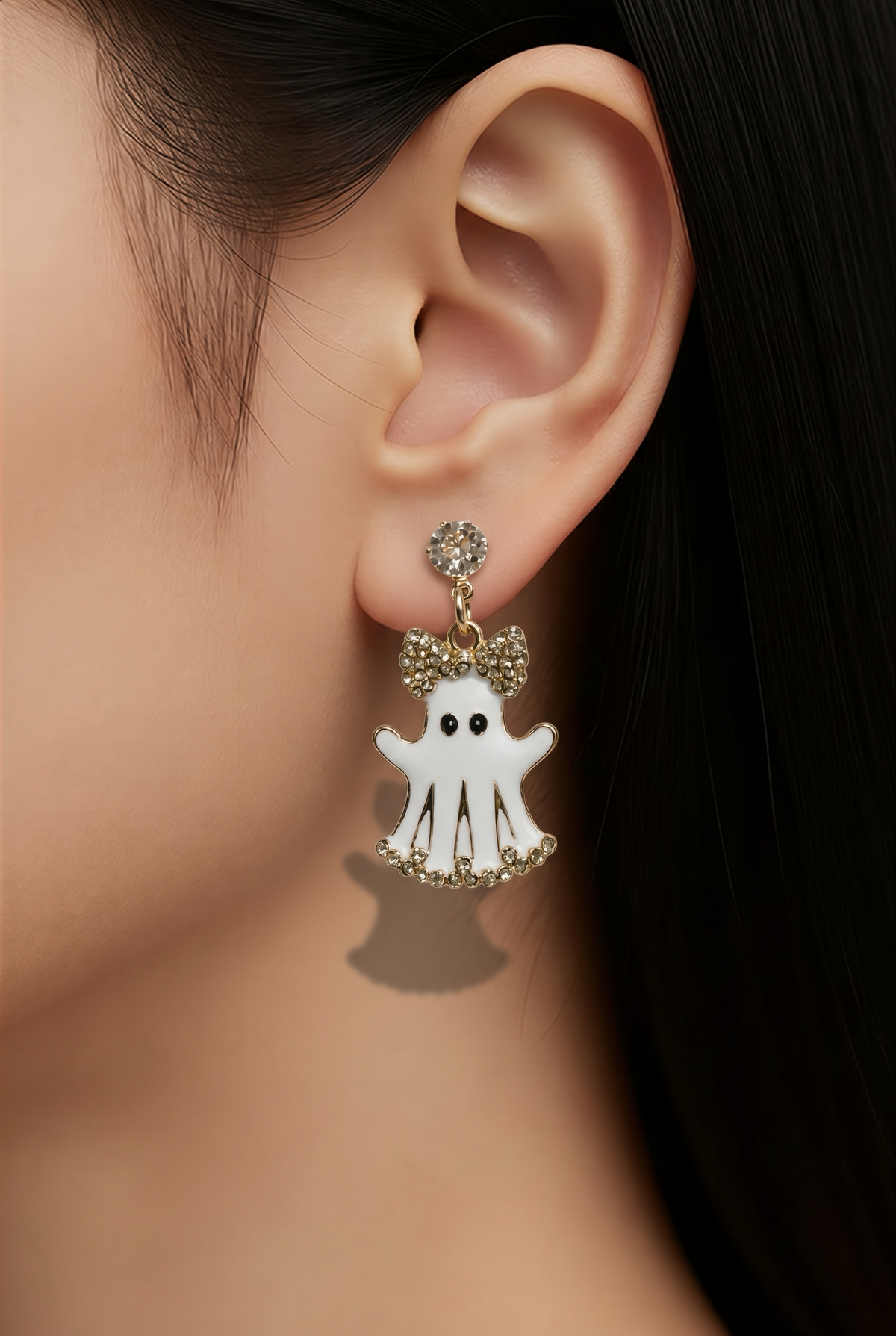 A close-up of a model's ear wearing a dangling earring with a white ghost charm, gold-tone bow details, and small clear rhinestones.