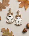 A pair of white ghost-shaped dangling earrings with gold-tone trim, rhinestone bow details, and small black eyes, styled on a textured beige fabric with autumn leaves and acorns.