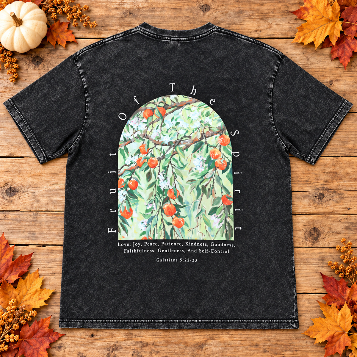 A black washed graphic t-shirt laid flat on a rustic wooden background decorated with autumn leaves and a small pumpkin. The t-shirt features a large arched graphic in the center showing an olive tree with red olives and green leaves, surrounded by the words 'Fruit of the Spirit' and a list of biblical virtues from Galatians 5:16-18.