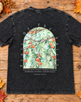 A black washed graphic t-shirt laid flat on a rustic wooden background decorated with autumn leaves and a small pumpkin. The t-shirt features a large arched graphic in the center showing an olive tree with red olives and green leaves, surrounded by the words 'Fruit of the Spirit' and a list of biblical virtues from Galatians 5:16-18.
