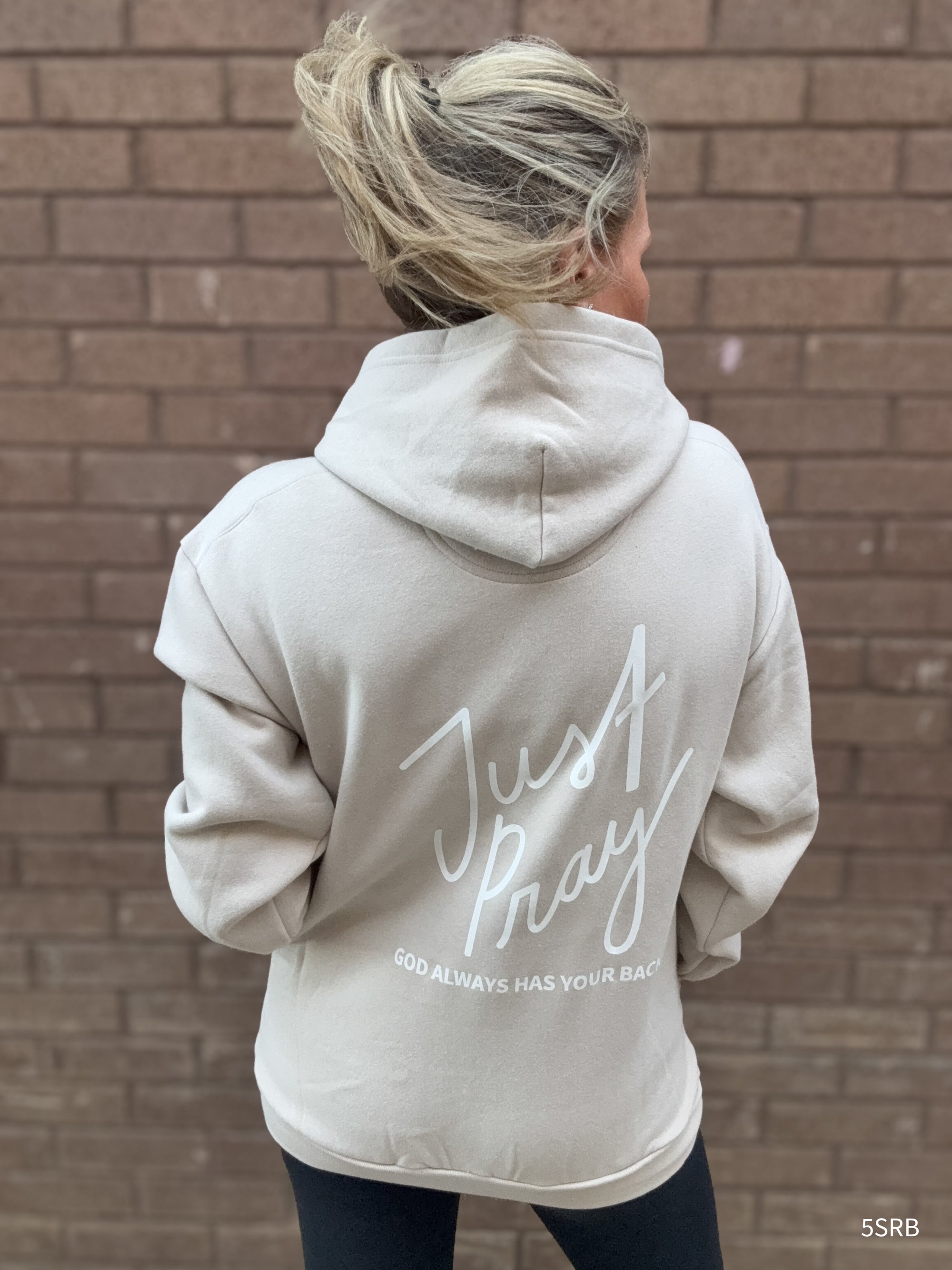 Person wearing a beige hoodie with 'Just Pray' text on the back against a brick wall.