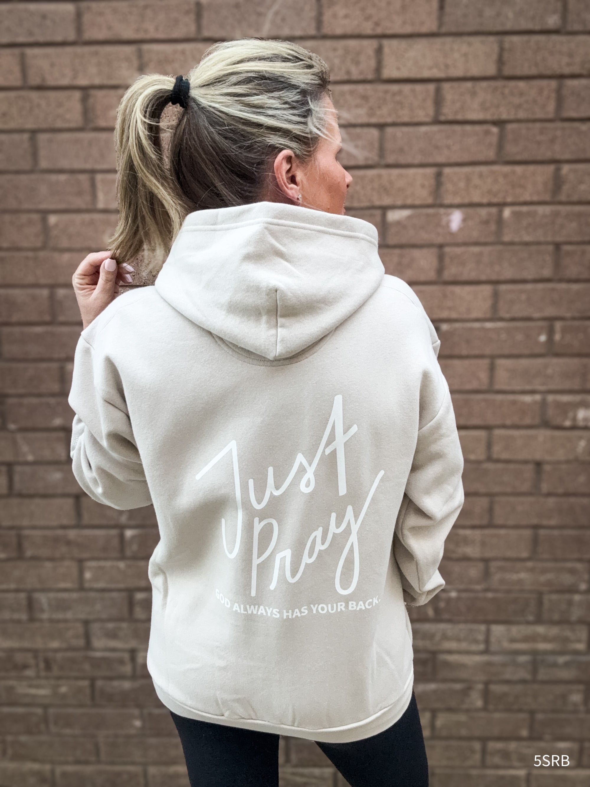 Person wearing a beige hoodie with 'Just Pray' text on the back against a brick wall.