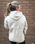 Person wearing a beige hoodie with 'Just Pray' text on the back against a brick wall.
