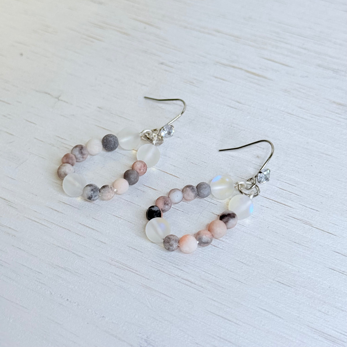 A pair of drop earrings with irregular polished beads in pink, gray, white, and black, suspended from silver fishhook wires with small clear rhinestone accents, shown on a white background.