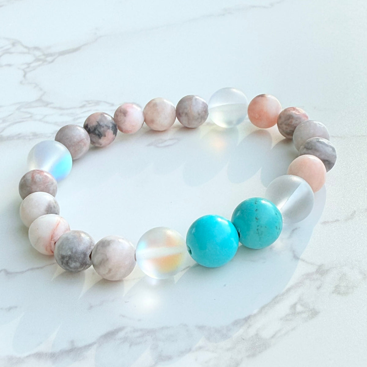 A bracelet with round beads in shades of turquoise, pink, white, and gray, displayed on a marble surface.