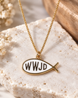 A gold fish-shaped pendant necklace with a white enamel center and the black letters 'WWJD' inside, displayed on a textured stone slab with small white flowers.
