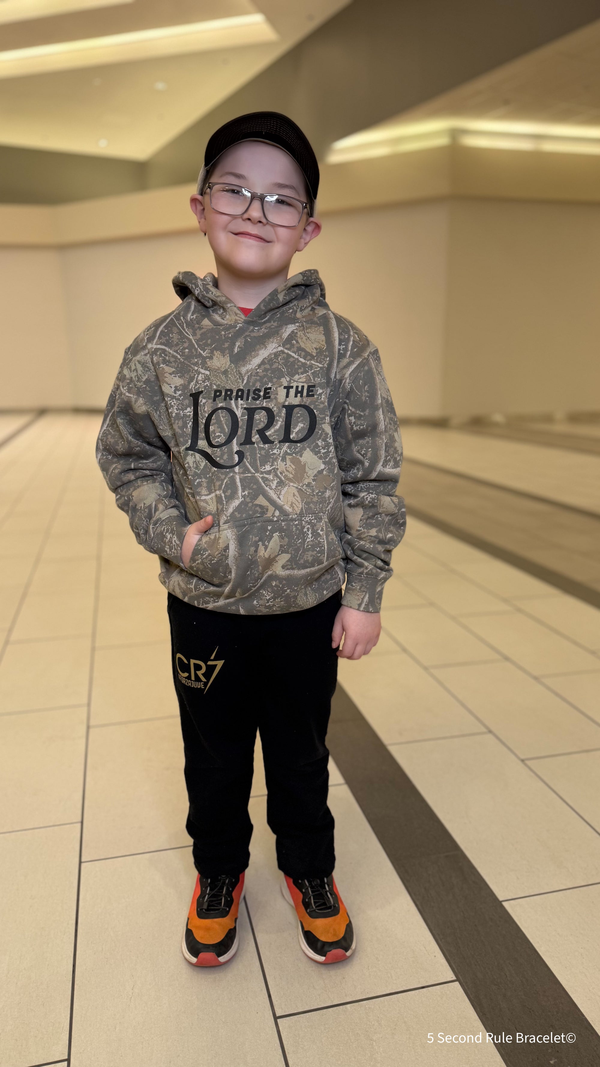 Child wearing a camouflage hoodie with text, standing in an indoor setting.