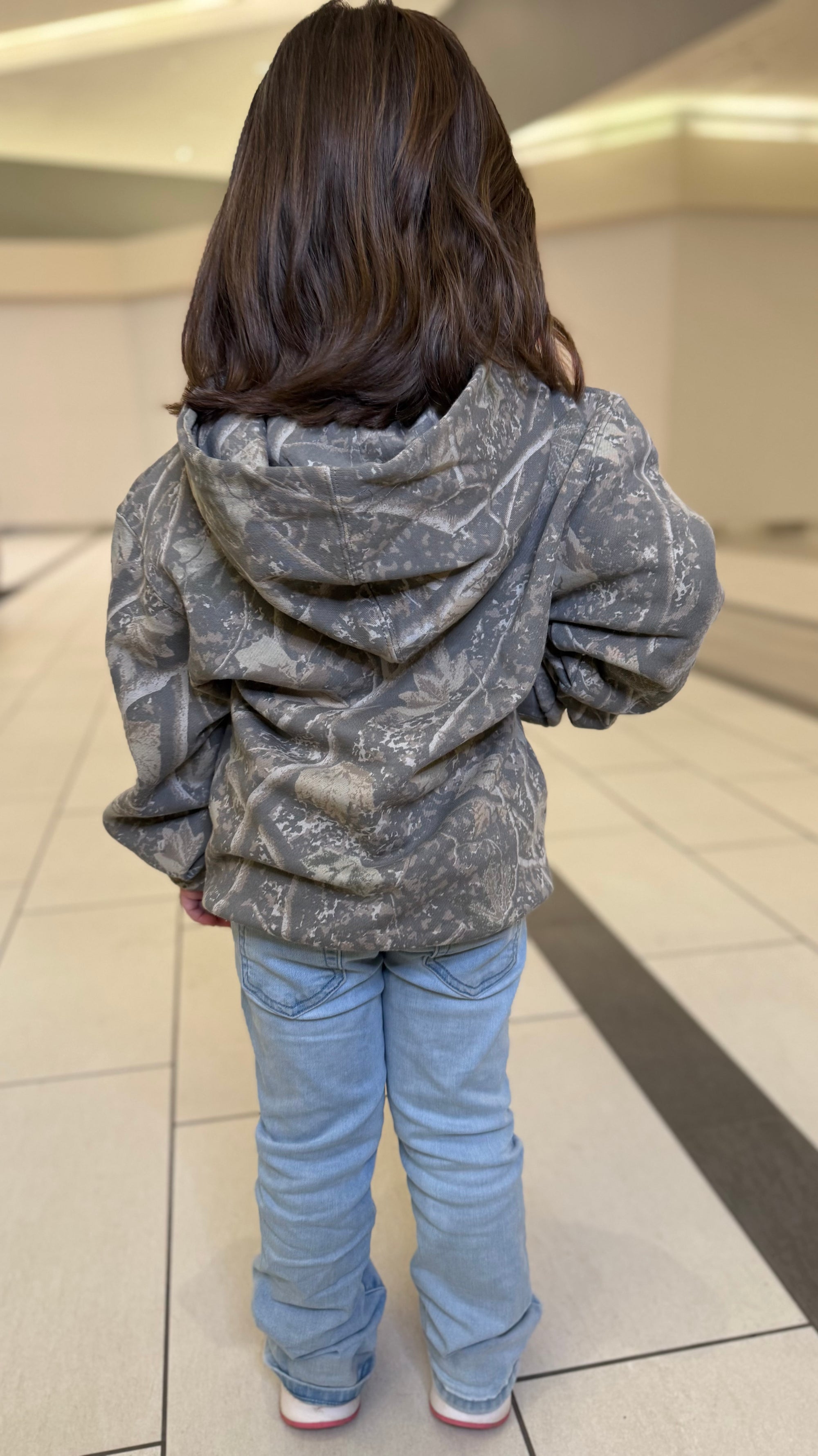 Child wearing a camouflage hoodie and blue jeans standing on a tiled floor.