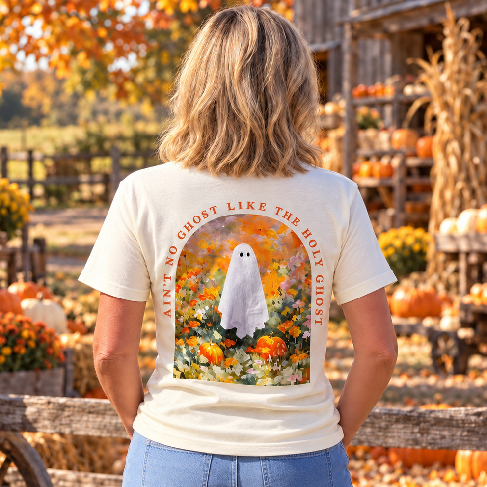 A woman stands in a pumpkin patch wearing a cream-colored graphic t-shirt. The back of the shirt features a large graphic of a white ghost surrounded by orange pumpkins and marigolds, with the text 'AIN'T NO GHOST LIKE THE HOLY GHOST' arched above the design.