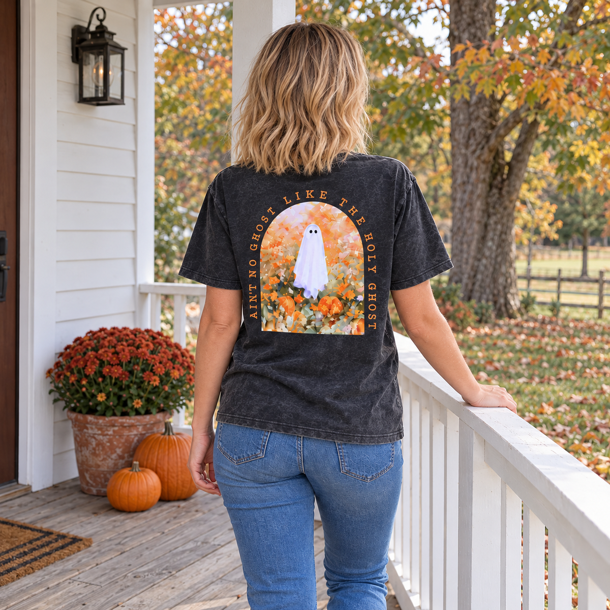 A woman stands on a white porch wearing a faded black graphic T-shirt from the back, showing a large design of a ghost in a field with autumn leaves and the text 'ain't NO GHOST LIKE THE holy GHOST ' She is also wearing blue jeans, with pumpkins and orange flowers on the porch beside her.