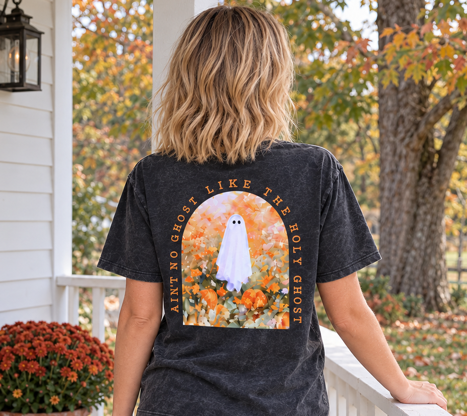 A woman stands on a white porch wearing a faded black graphic T-shirt from the back, showing a large design of a ghost in a field with autumn leaves and the text 'ain't NO GHOST LIKE THE holy GHOST ' She is also wearing blue jeans, with pumpkins and orange flowers on the porch beside her.