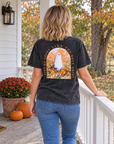 A woman stands on a white porch wearing a faded black graphic T-shirt from the back, showing a large design of a ghost in a field with autumn leaves and the text 'ain't NO GHOST LIKE THE holy GHOST ' She is also wearing blue jeans, with pumpkins and orange flowers on the porch beside her.