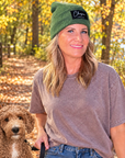 A woman wearing a green beanie with a black 'Chicory Farms' logo and a faded brown t-shirt stands outdoors on a leaf-covered path with a brown dog beside her.