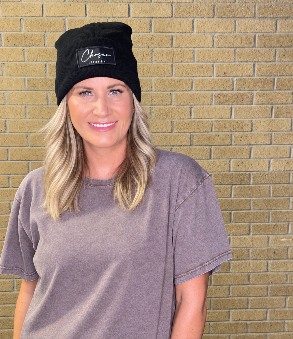 A woman with blonde hair wearing a black beanie with a white embroidered logo and a faded gray t-shirt, standing in front of a tan brick wall.