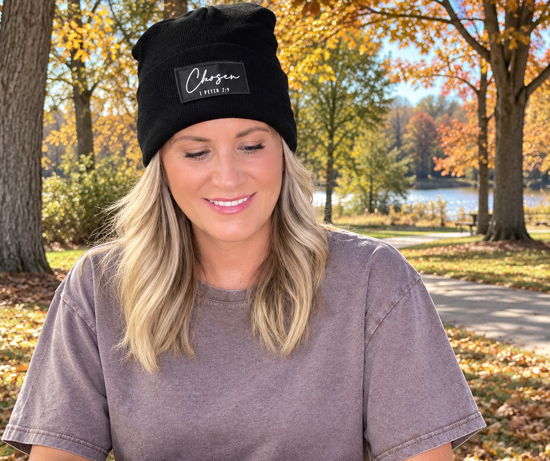 A woman sitting at a wooden table outdoors in a park, wearing a black beanie with a white 'Chosen' patch and Trees with autumn foliage and a lake are visible in the background.