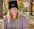 A woman sitting at a wooden table outdoors in a park, wearing a black beanie with a white 'Chosen' patch and Trees with autumn foliage and a lake are visible in the background.