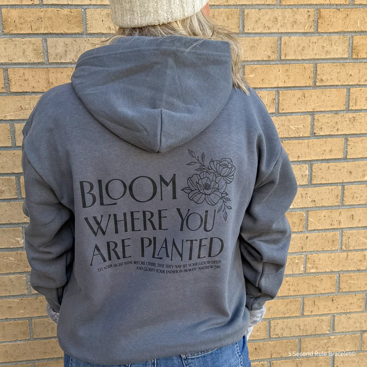 Person wearing a gray hoodie with 'Bloom Where You Are Planted' text and floral design on the back, standing against a brick wall.