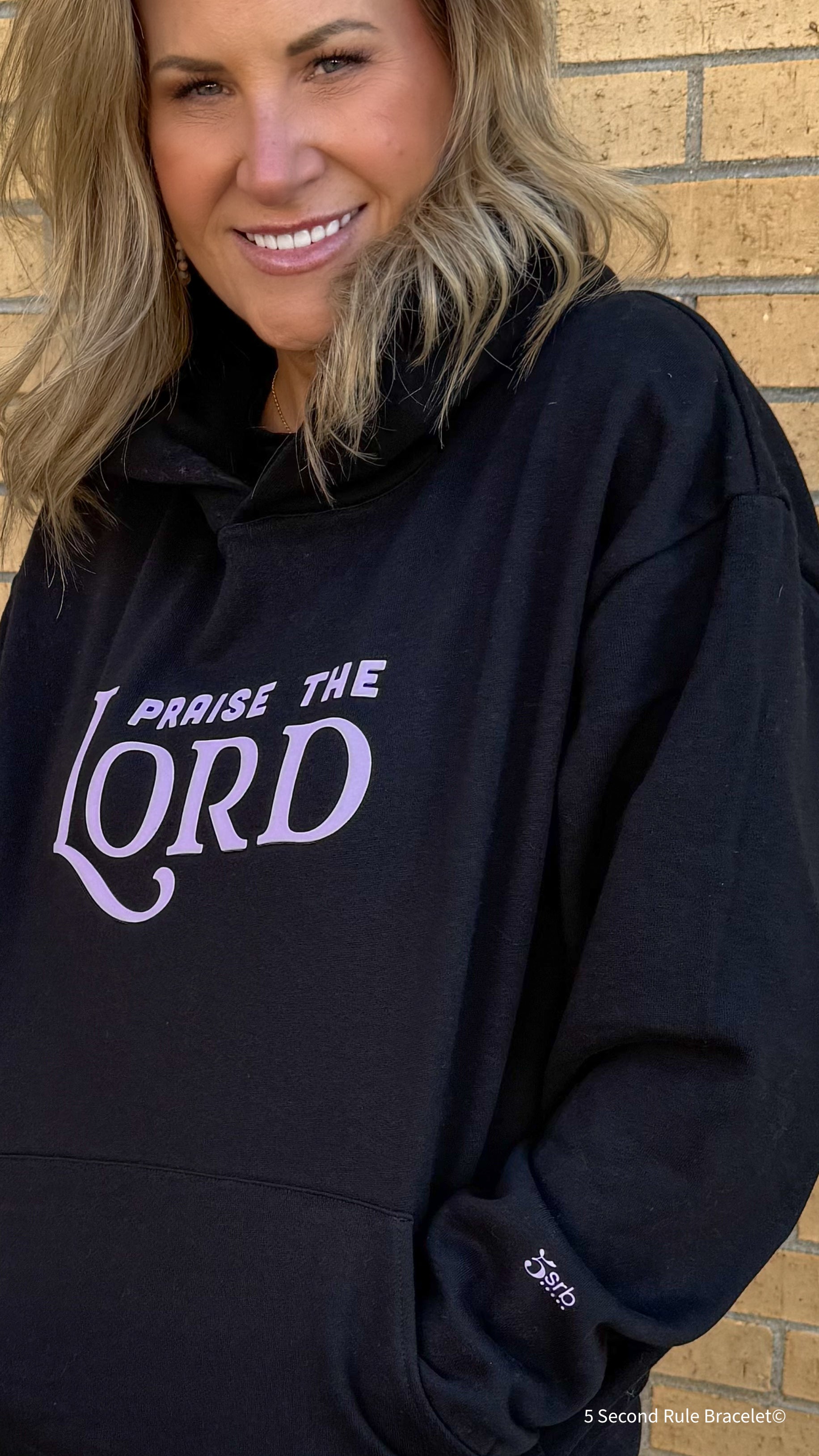 Person wearing a black hoodie with 'Praise the Lord' text against a brick wall.