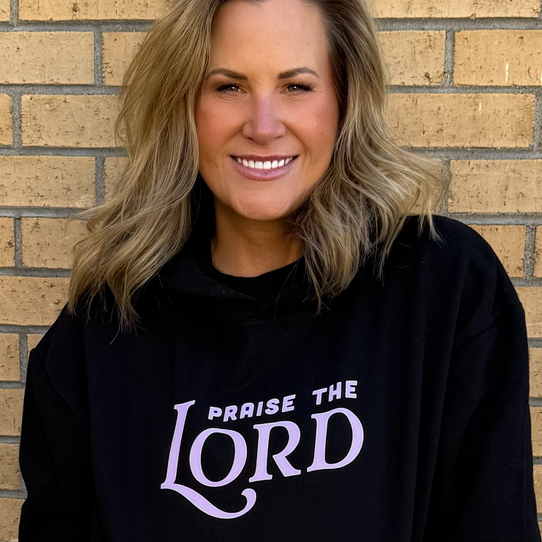 Woman wearing a black hoodie with 'Praise the Lord' text against a brick wall.